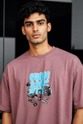 A man wearing a dusty rose crewneck t-shirt with a colorful graphic print that reads 'ELSH BANK' in light blue, featuring cartoon speakers, a boombox, and other retro electronic accessories.