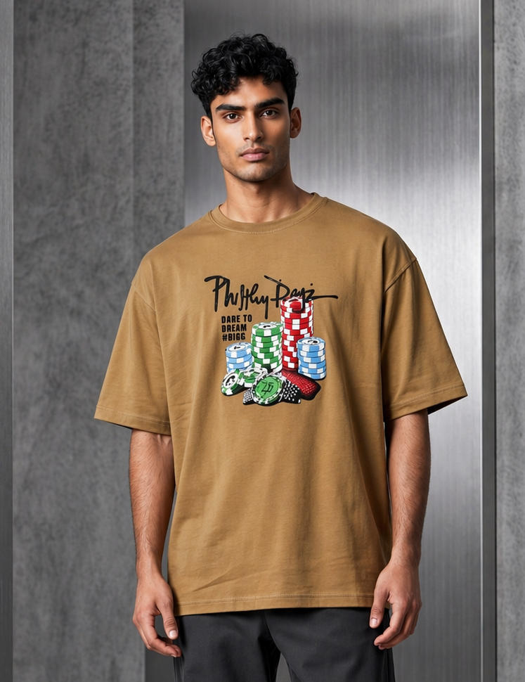 A man modeling an oversized tan graphic t-shirt with a colorful hand-drawn design and text, paired with dark gray trousers and white sneakers, standing against a gray concrete wall.